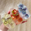 A hand holds three clear glass bottles filled with colored liquid and fruit pieces, resembling a fruit bottle stress ball set