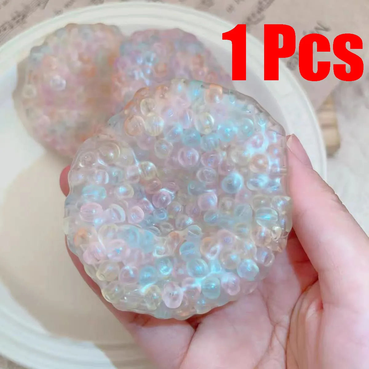 A hand holds a clear, iridescent squishy stress relief ball with pink and blue hues on a white plate.