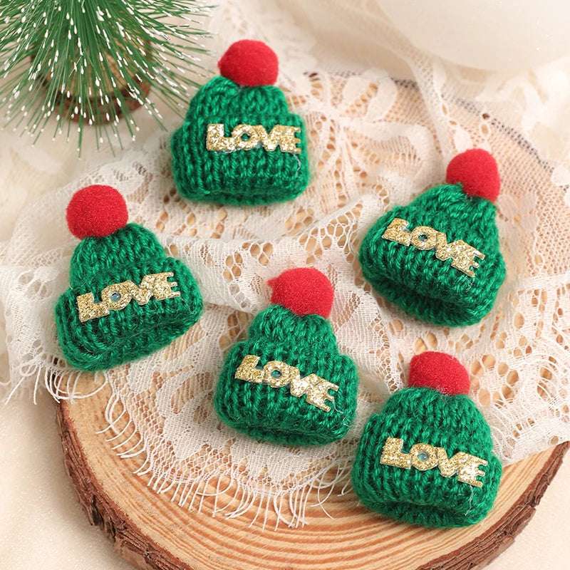 A set of mini knitted green hats with red pom-poms and gold LOVE embroidery displayed on a wooden surface.