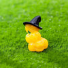 A small black witch hat figurine sits atop a bright yellow pumpkin bear figurine on green grass.