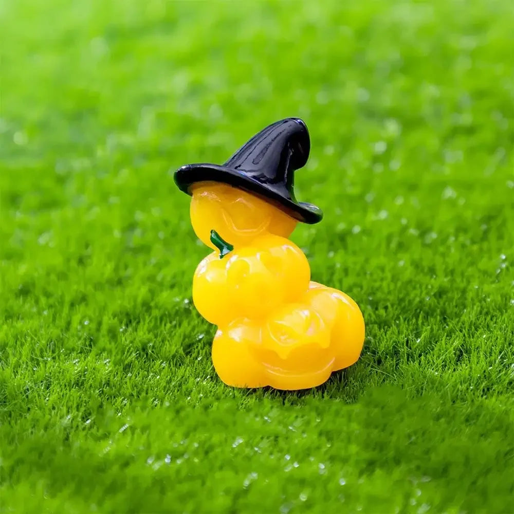 A small black witch hat figurine sits atop a bright yellow pumpkin bear figurine on green grass.