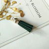 A silver tassel with a dark green velvet cord rests on a white card next to dried yellow floral elements.