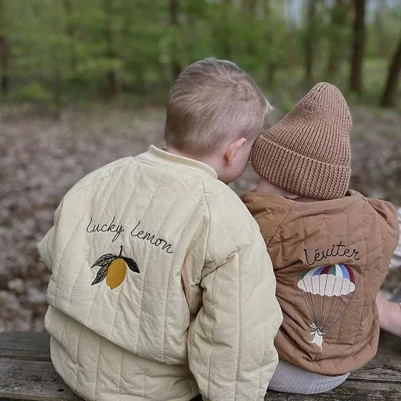 A young child wearing a beige padded jacket with a letter print sitting outdoors.