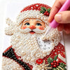 A hand applies pearl art to a Christmas ornament featuring a bearded man and festive red and green details.