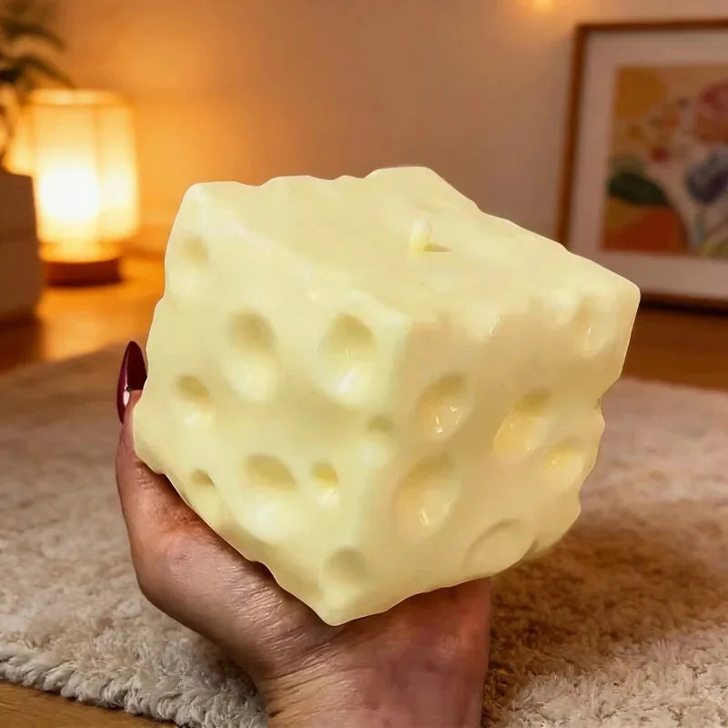 A hand holds a large, pale yellow cheese-shaped squishy fidget toy on a beige surface.