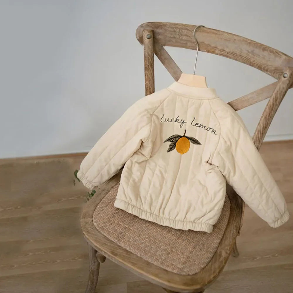 A cream padded jacket for kids with a yellow lemon letter print displayed on a wooden chair.