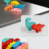 Une main pose une pièce de dinosaure en bois rouge sur une pile de dinosaures colorés sur une table grise.