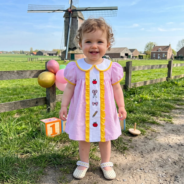 Kleines Mädchen in einem rosa karierten Kleid mit gelbem Zierrand und Stickerei in einer Wiese.