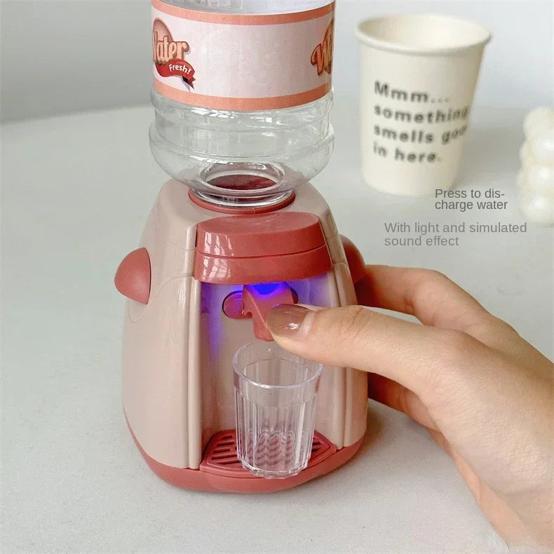 A hand is pressing a pink coffee maker water dispenser toy, which has a clear water cup and a label reading Water Fresh.