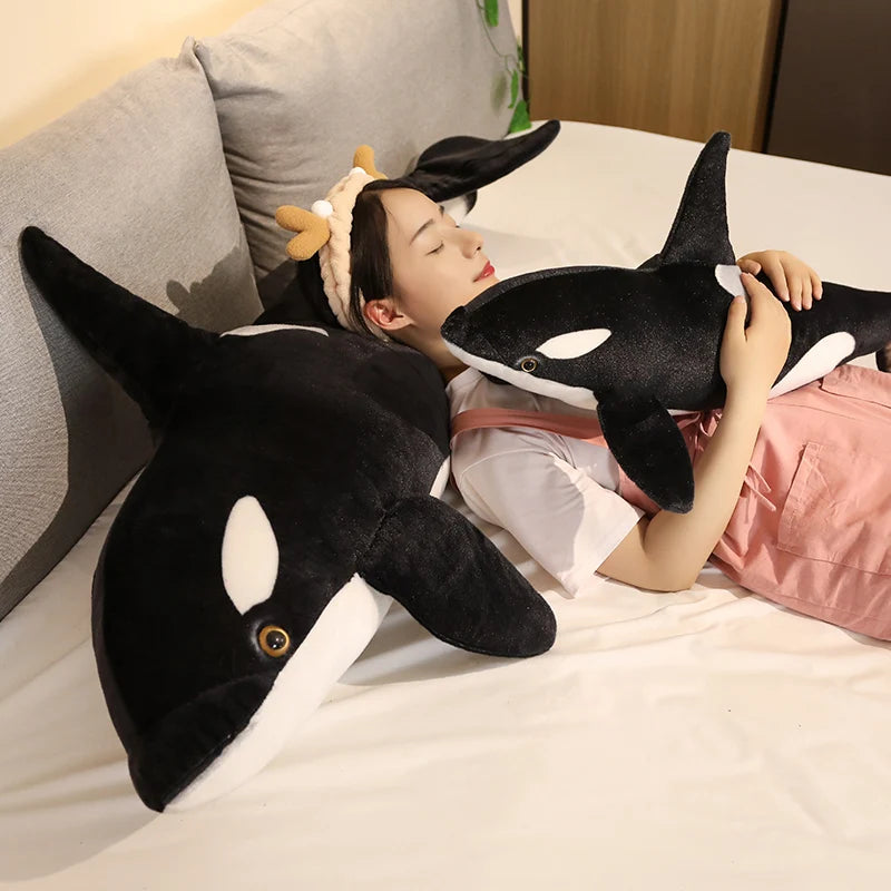 A woman is lying down with a black and white orca whale plush toy on her chest in a bright bedroom setting.