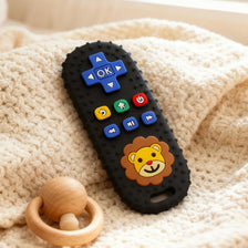 A black silicone teether shaped like a lion's face with various buttons and a remote control design resting on a cream-colore