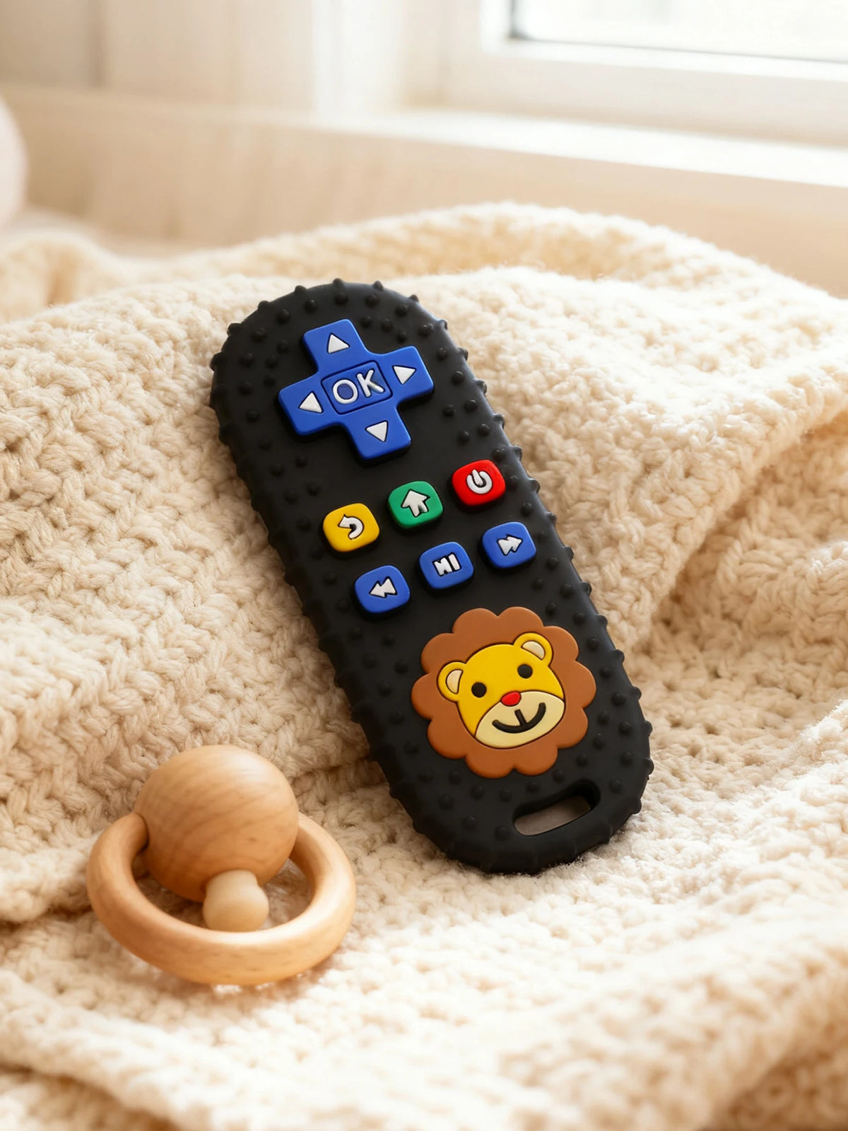 A black silicone teether shaped like a lion's face with various buttons and a remote control design resting on a cream-colore