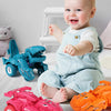 A smiling baby is sitting on the floor surrounded by various pink and blue dinosaur transforming car toys.