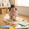A young girl holds a handmade crochet sloth and squirrel plush toy on a fluffy white rug surrounded by wooden blocks.