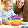 A toddler sits next to an adult while playing with a stack of colorful plastic cups and boats.