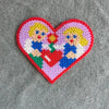 A heart-shaped mosaic created from colorful fuse beads depicting two stylized female figures.