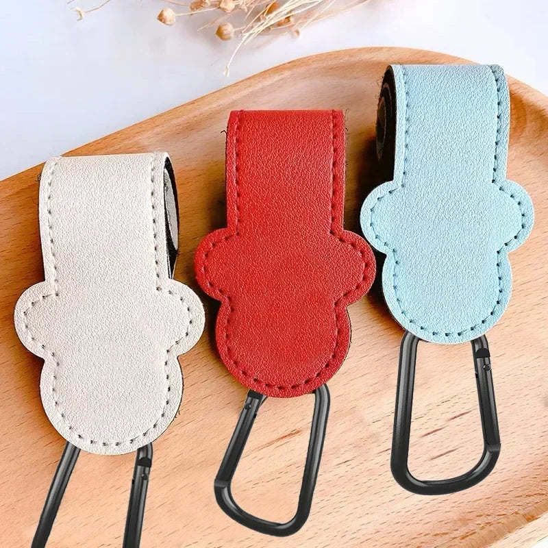 Three colored PU leather stroller organizer hooks in beige, red, and light blue displayed on a wooden board.