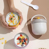 A hand holds a yellow bowl of breakfast cereal and a white spoon next to a gray insulated stainless steel cup with a lid.