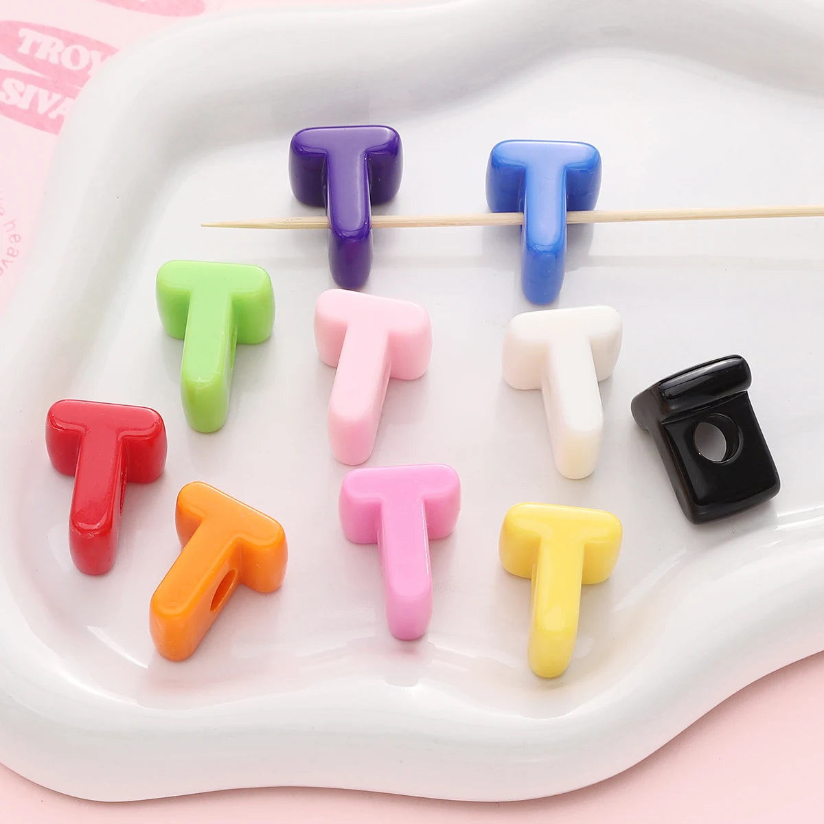 A collection of colorful acrylic letter beads shaped like the letter T in various shades and sizes arranged on a white tray.