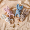A set of handmade crochet bunny rattles in pink, blue, and cream colors resting on a light fabric.