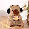 A soft brown capybara plush toy is sitting on a wooden surface and holding a pink lotus flower.