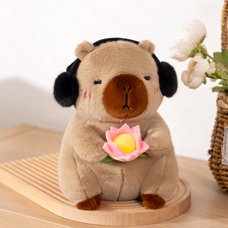 A soft brown capybara plush toy is sitting on a wooden surface and holding a pink lotus flower.
