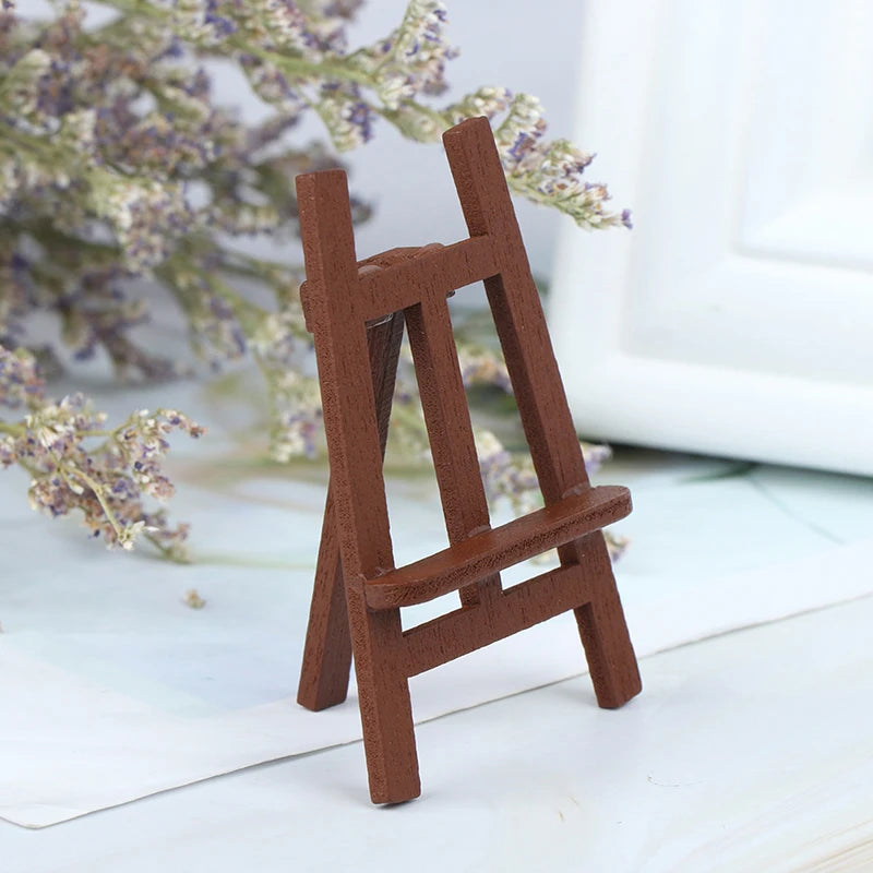 A small, rustic wooden artist easel used for dollhouses, resting on a white surface with lavender branches in the background.