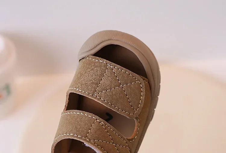 A close-up of a tan suede toddler sandal for children, showing the stitching and soft sole.