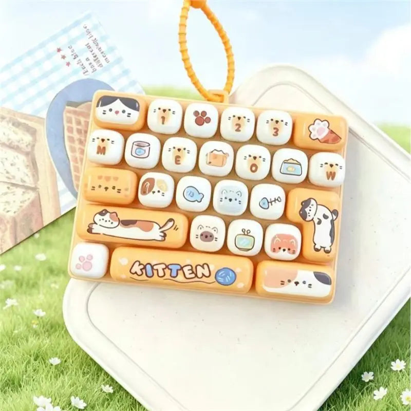 A set of 26 cartoon animal keycap charms arranged in a rectangular tray, displayed on a white surface with green grass in the