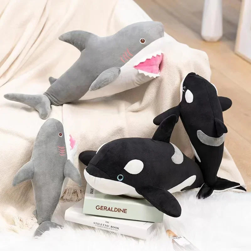 A collection of gray and black plush stuffed animals depicting a killer whale and a great white shark resting on a light fabr