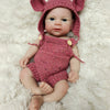 A realistic silicone reborn doll with pink ears and a matching knitted outfit lying on white fluffy fur.