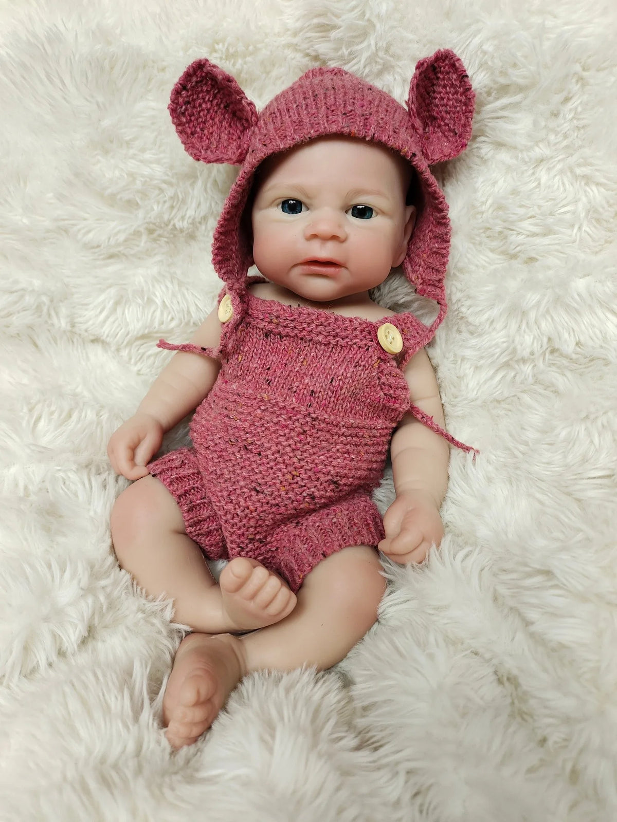 A realistic silicone reborn doll with pink ears and a matching knitted outfit lying on white fluffy fur.