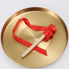 A polished brass alloy gong with a wooden mallet tied with a red ribbon for meditation or ceremonial use.