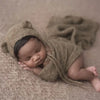 A newborn baby is lying on a beige surface wearing a knitted bear hat and wrap set.