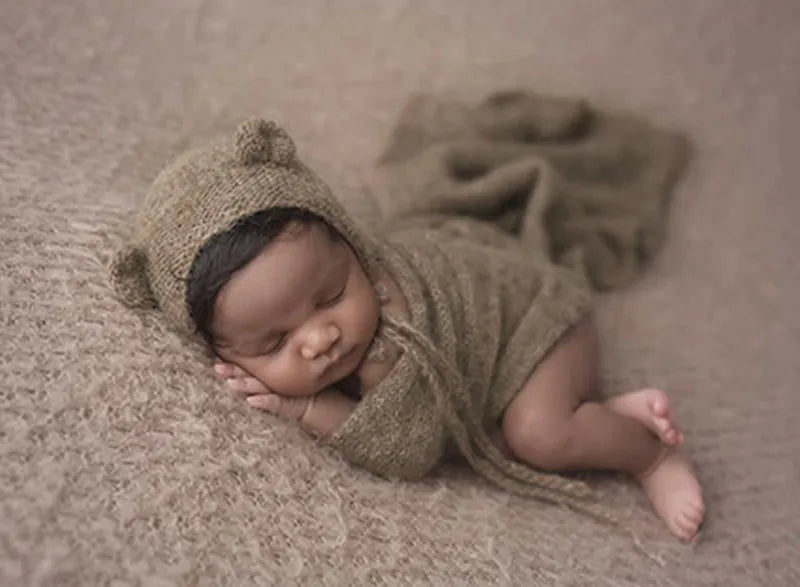 A newborn baby is lying on a beige surface wearing a knitted bear hat and wrap set.