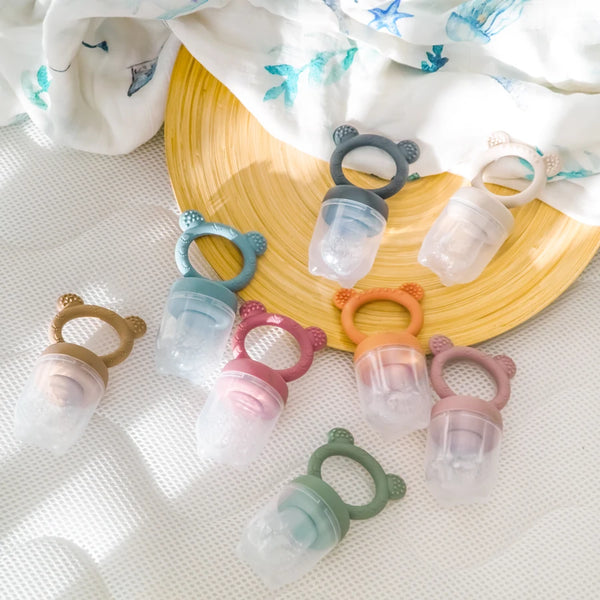 A collection of silicone fruit feeder pacifiers in various colors and shapes arranged on a yellow wooden plate.
