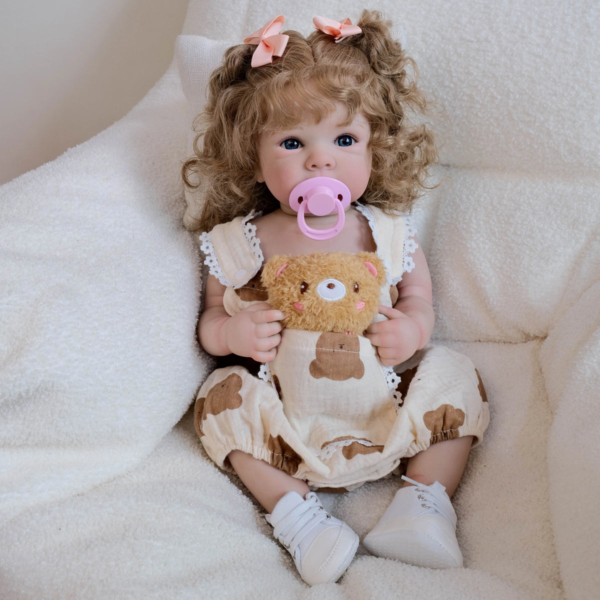 A lifelike 18-inch baby doll with hand-rooted hair wearing a light brown dress and pink pacifier, sitting in white fluffy fab