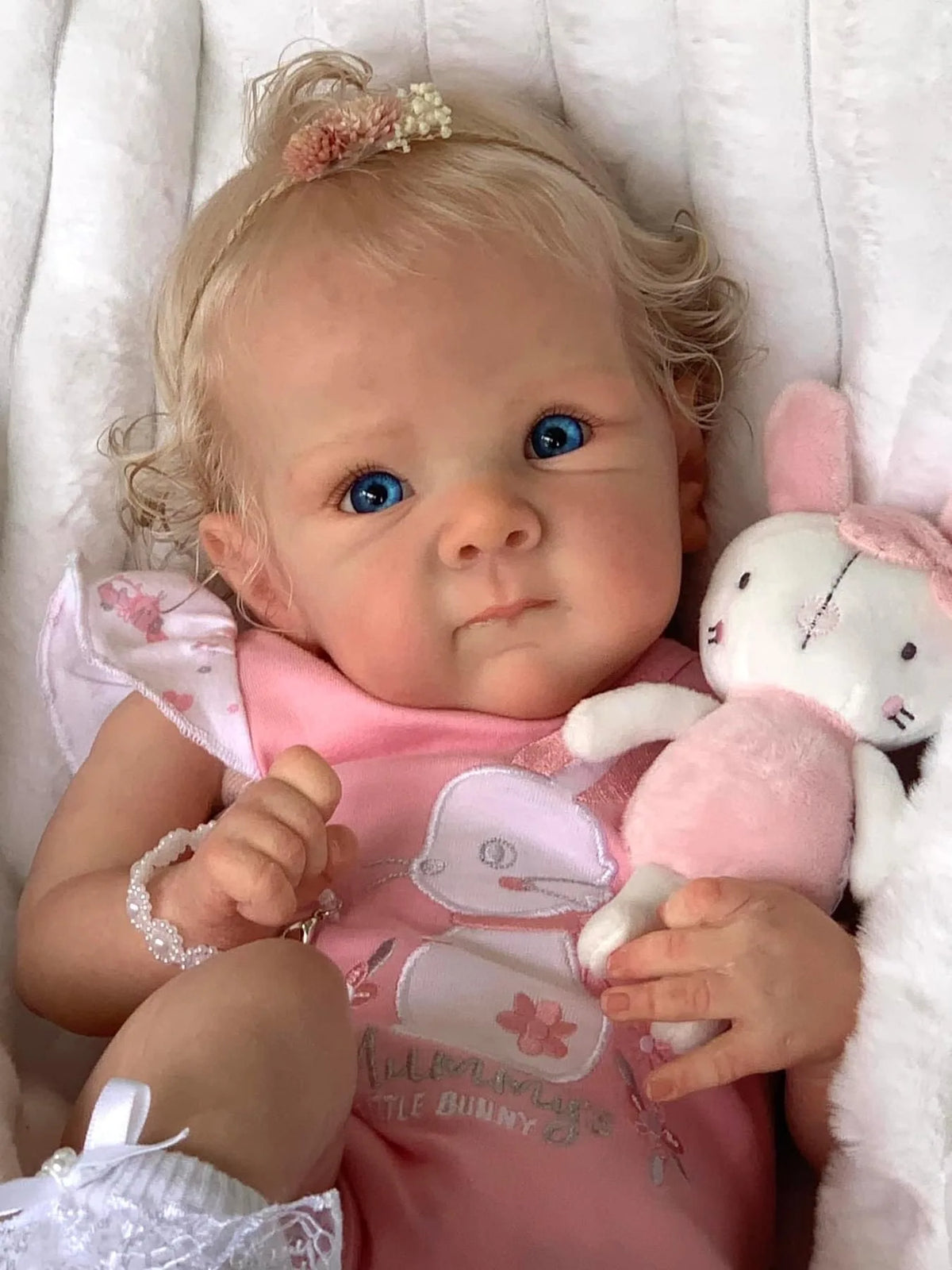 A close-up photo of a lifelike reborn baby doll with hand-rooted hair, featuring blue eyes and pink clothing.