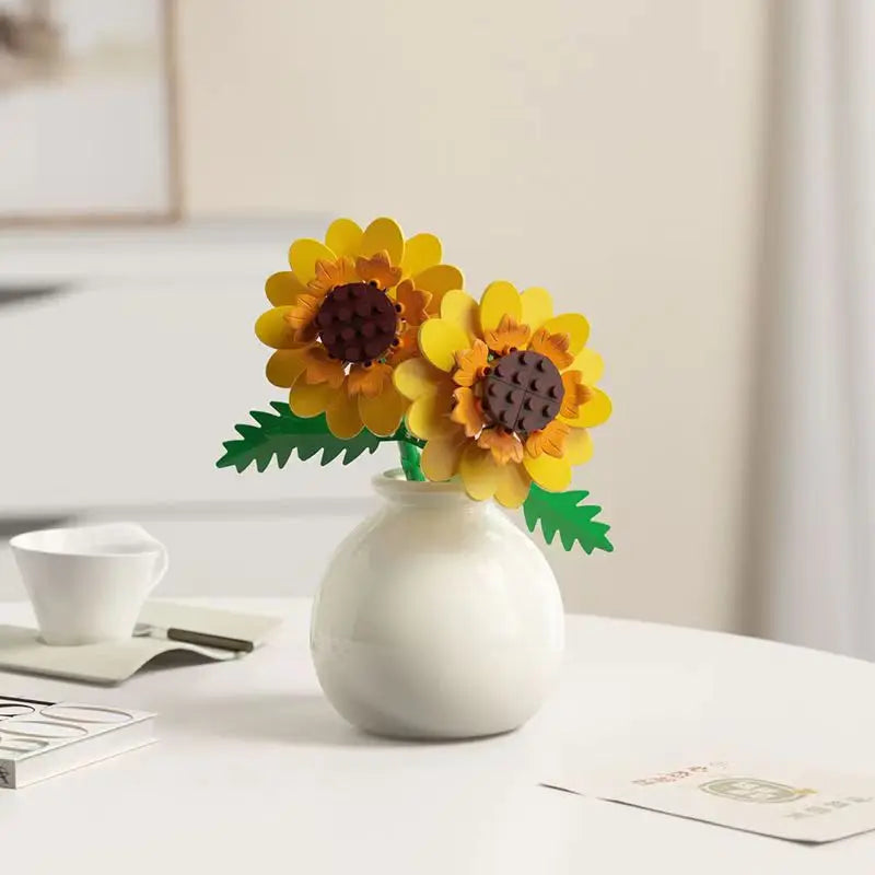 A white ceramic vase holds a bouquet of yellow and brown flowers with green leaves on a white table.