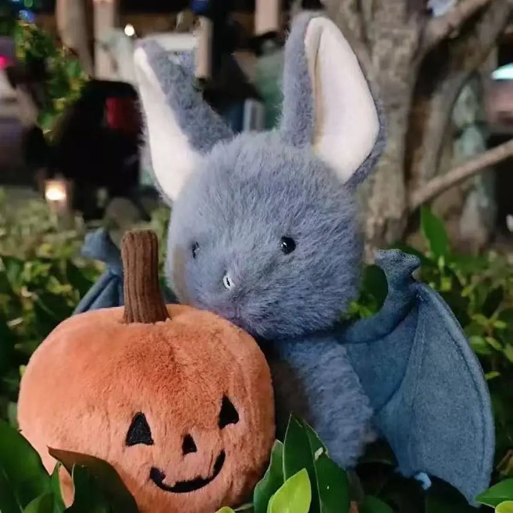 A soft grey bat plush toy with large ears and blue wings is resting near a pumpkin-shaped item.
