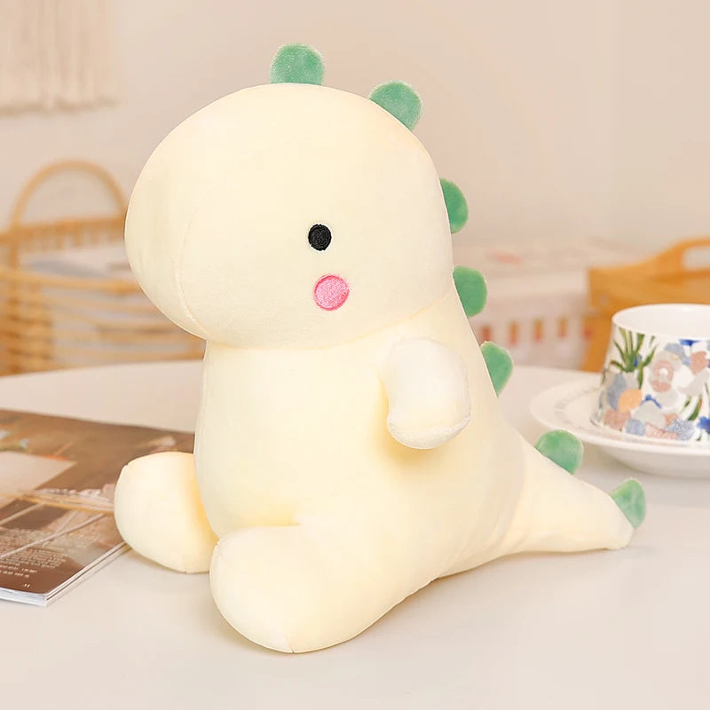 A soft yellow dinosaur plush toy with green spikes sitting on a white surface.