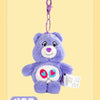 A purple Care Bears plush toy keychain with a pink charm attached, displayed against a light yellow background.