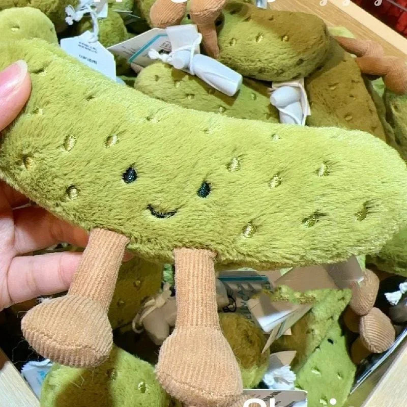 A soft green plush pickle toy with a smiling face is shown in a box.