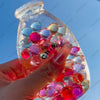 A hand holds a clear bottle filled with colorful, squishy fruit-shaped stress balls.
