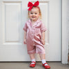 A smiling toddler girl wearing a pink satin button-up romper sleepwear with a red bow.