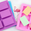 A pink silicone soap mold with six square cavities and a light green mold next to a pink flamingo illustration.