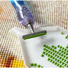 A purple diamond painting pen is shown applying green beads onto a white tray on a textured fabric background.