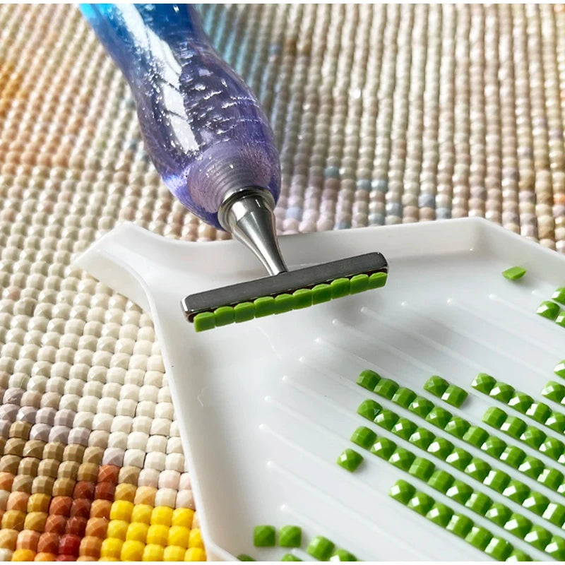 A purple diamond painting pen is shown applying green beads onto a white tray on a textured fabric background.