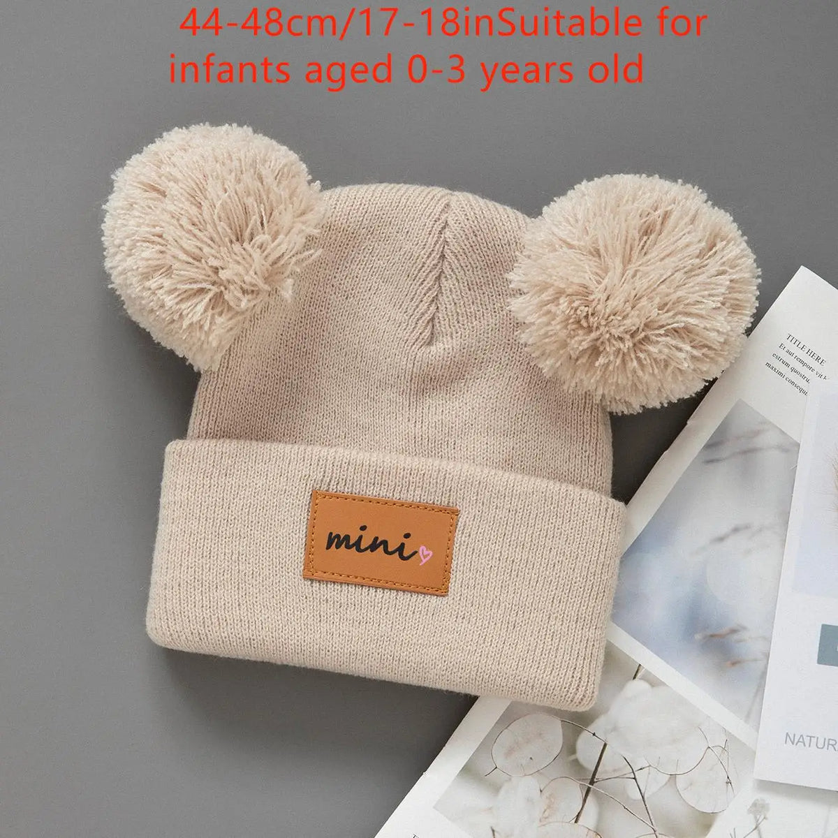 A beige knit baby beanie hat with fluffy pom-poms, suitable for infants aged 0-3 years old.