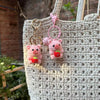 A handmade crochet mini pig keychain with pink ears and a red nose hanging from a gold-colored ring on a beige woven bag.